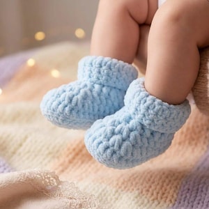 Puede incluir: Un par de botines de bebé de punto azul claro. Los botines están hechos de un hilo suave y texturizado y tienen un puño enrollado. Los botines de bebé están sobre una manta colorida con una textura suave y difusa. Los botines son una prenda para bebé.