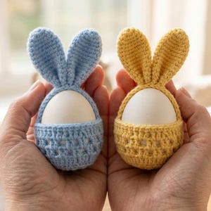 May include: Two handmade crochet egg holders, one light blue and one yellow, each shaped like a bunny with long ears. The egg holders are designed to cradle a white egg, perfect for Easter.