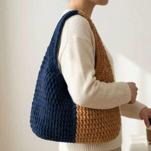 May include: A handmade, two-tone crochet shoulder bag. The bag features a deep navy blue section and a tan section, with a matching navy blue strap. The bag is made of a thick, textured yarn, and is designed to be worn over the shoulder.