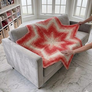 May include: A star-shaped, hand-crocheted blanket in shades of red and white rests on a gray sofa. The blanket's design features a central star pattern with radiating points. The yarn appears soft and textured, perfect for home decor.