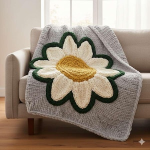 May include: A gray knitted blanket with a large daisy design. The daisy has a yellow center, white petals, and a green border. The blanket is draped over a beige sofa, creating a cozy and inviting aesthetic.