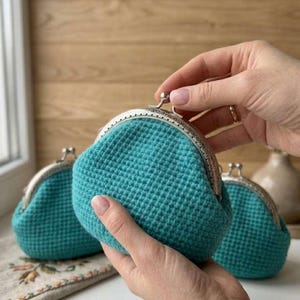 Könnte beinhalten: Türkisfarbene Häkeltasche mit silbernem Verschluss. Die Tasche hat eine strukturierte, abgerundete Form und wird von den Händen einer Person gehalten. Zwei weitere Taschen sind im Hintergrund zu sehen. Die Taschen sind aus weichem, strukturiertem Garn gefertigt.