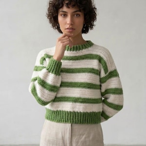May include: A cream-coloured, crocheted sweater with horizontal green stripes. The sweater has a crew neck and long sleeves with green cuffs. The model is wearing the sweater with beige trousers.