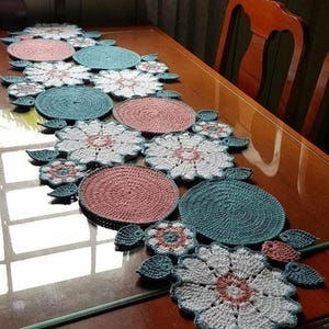 Può includere: Runner da tavolo all'uncinetto fatto a mano con un motivo floreale. Il runner presenta elementi circolari e a forma di fiore alternati in rosa, verde acqua e bianco, con accenti di foglie verdi. Su un tavolo di legno.