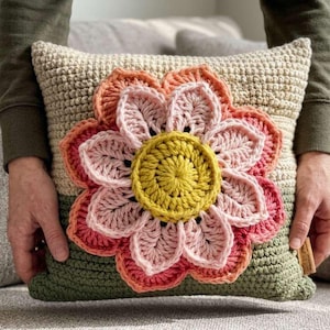 May include: A crocheted pillow with a large, colorful flower design. The flower features petals in shades of pink and orange, with a yellow center. The pillow has a beige background and a green base.