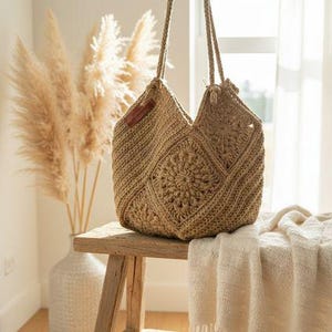 Puede incluir: Un bolso tote de crochet beige hecho a mano con asas de cuerda. El bolso presenta un diseño geométrico con un motivo floral central. Descansa sobre un taburete de madera rústica, con una manta color crema cerca. El fondo incluye hierba de la pampa en un jarrón blanco.