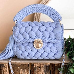 Puede incluir: Un bolso de mano de ganchillo azul claro hecho a mano con un diseño trenzado. El bolso presenta un asa superior, un cierre de solapa con un cierre dorado y una borla larga. También es visible una correa de cadena plateada.