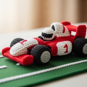 Puede incluir: Un coche de carreras rojo y blanco hecho a mano con ganchillo, con ruedas negras y el número 1. El coche de juguete está colocado sobre una pista de fieltro verde con líneas blancas. El coche mide unos 15 cm de largo.