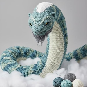 May include: A crocheted sea serpent in shades of blue, green, and white, with yellow eyes and a gray beard. The serpent is posed on a bed of fluffy white material, with yarn and a crochet hook nearby.