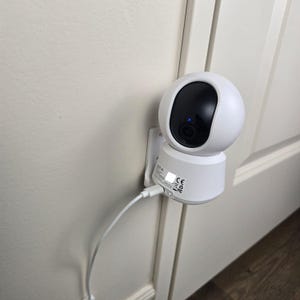 May include: A white spherical security camera with a black lens housing and a small blue indicator light. The device is mounted to a wall and connected to a white power cable. The base of the camera features printed technical text and icons.