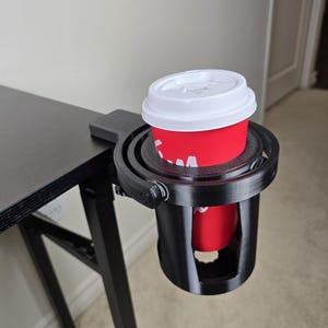 May include: A black, 3D-printed cup holder clamped to a black table. A red coffee cup with a white lid is held securely. The holder is designed to attach to the edge of a table, providing a convenient spot for drinks.