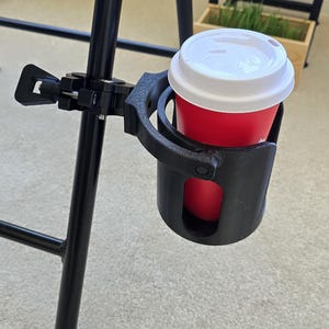 May include: A black plastic cup holder attached to the metal leg of a folding chair. The holder features a clamp mechanism with a tightening knob and contains a red disposable cup with a white plastic lid, securely held in the circular frame.