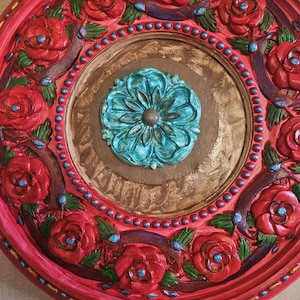 May include: A round, decorative wall art piece with a vibrant red border. The design features sculpted red roses, teal accents, and a central teal floral medallion. The inner circle has a textured brown finish.