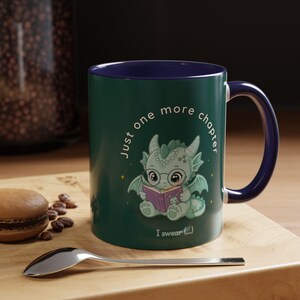 Könnte beinhalten: Eine grüne Keramiktasse mit blauem Innenraum und Henkel. Die Tasse zeigt einen Cartoon-Drachen, der ein Buch liest, mit dem Text "Just one more chapter, I swear (...)". Die Tasse steht auf einer Holzoberfläche mit einem Löffel und einem Gebäck.