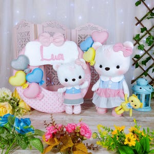 May include: Two white teddy bear plush toys, one large and one small, stand in front of a pink decorative backdrop. The large bear wears a blue and pink dress and holds a yellow bear. The small bear wears a similar dress. A pink wreath with the name "Lua" is in the background.