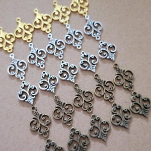 May include: A collection of metal fleur-de-lis charms arranged in rows. The charms feature intricate scrollwork and are finished in gold, silver, and antique bronze tones. Each piece includes a small loop at the top for attachment to jewellery.