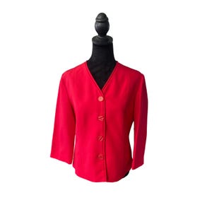 Könnte beinhalten: Ein leuchtend roter Blazer mit V-Ausschnitt und drei passenden Knöpfen. Der Blazer hat Dreiviertelärmel und wird auf einer schwarzen Schaufensterpuppe präsentiert. Der Stoff hat eine glatte, einfarbige Optik.