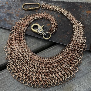 May include: A copper-coloured chainmail necklace with a matching chain and clasp. The necklace is made of metal rings, with a brass-coloured clasp and chain attachments. The chainmail is arranged in a wide, curved shape.