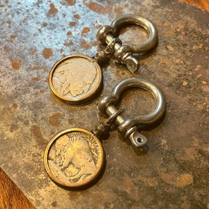 May include: Two silver keychains with a buffalo nickel design. Each keychain features a silver loop with a round buffalo nickel design. The buffalo nickel is a coin with a profile of a buffalo on one side and the head of an American Indian on the other side.