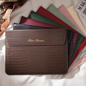 May include: A brown faux-leather laptop sleeve with a crocodile texture, featuring the name "Kaia Brown" in gold lettering. A fan of colorful laptop sleeves in various colors is displayed behind the brown sleeve. Accessories like a perfume bottle, sunglasses, and a magazine are also visible.