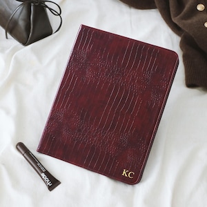 May include: A dark burgundy tablet case with a textured crocodile skin pattern and the gold embossed initials KC in the bottom right corner. The case is positioned alongside a dark brown handbag, a brown cardigan, and a small tube of Rhode lip tint.