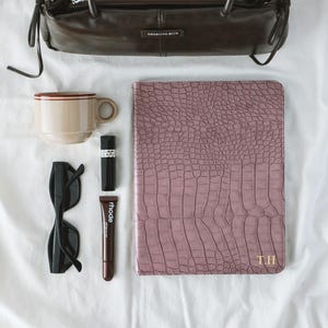 Puede incluir: Un bolso de cuero marrón oscuro con lazos laterales, una taza de cerámica beige con borde rojo, gafas de sol negras, un labial negro, un tubo de tratamiento labial Rhode marrón y un cuaderno malva con textura de cocodrilo y las iniciales doradas T.H. en la portada.