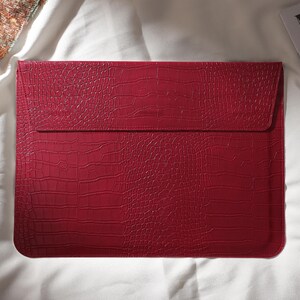 Puede incluir: Una funda roja para port&aacute;til con textura de piel de cocodrilo. La funda tiene un cierre de solapa y es de forma rectangular. La funda est&aacute; sobre una superficie blanca.