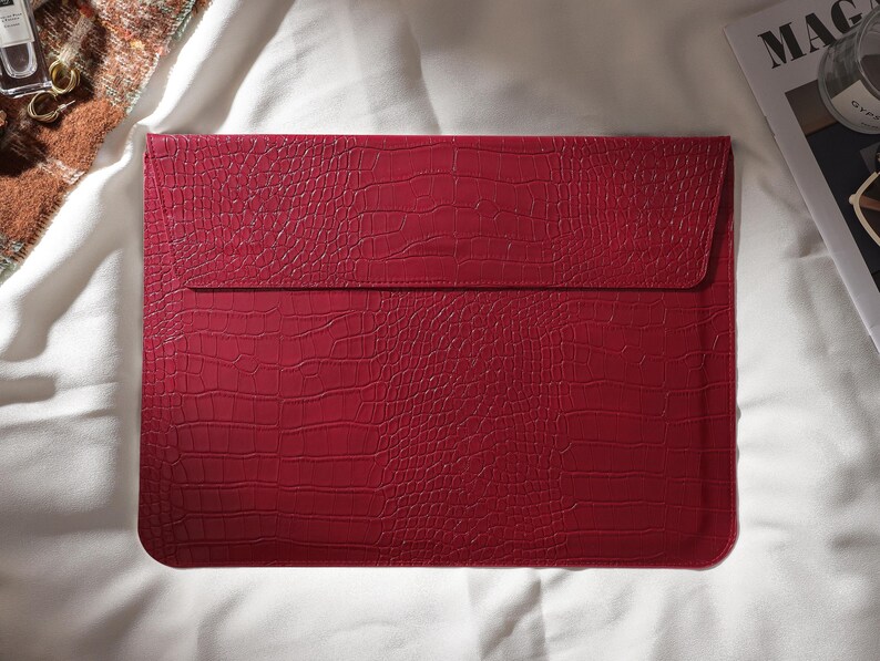 Puede incluir: Una funda roja para port&aacute;til con textura de piel de cocodrilo. La funda tiene un cierre de solapa y es de forma rectangular. La funda est&aacute; sobre una superficie blanca.