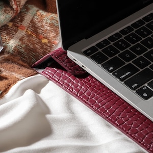 May include: A burgundy leather laptop sleeve with a textured, crocodile-like pattern. The sleeve is next to a silver laptop with a black keyboard. A perfume bottle, jewelry, and a patterned scarf are also visible.
