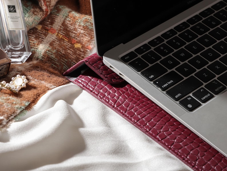 May include: A burgundy leather laptop sleeve with a textured, crocodile-like pattern. The sleeve is next to a silver laptop with a black keyboard. A perfume bottle, jewelry, and a patterned scarf are also visible.