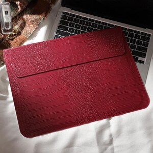 Puede incluir: Una funda roja para port&aacute;til con textura de piel de cocodrilo. La funda rectangular tiene un cierre de solapa. Est&aacute; sobre un teclado de port&aacute;til. El fondo incluye un frasco de perfume, un reloj y una manta.