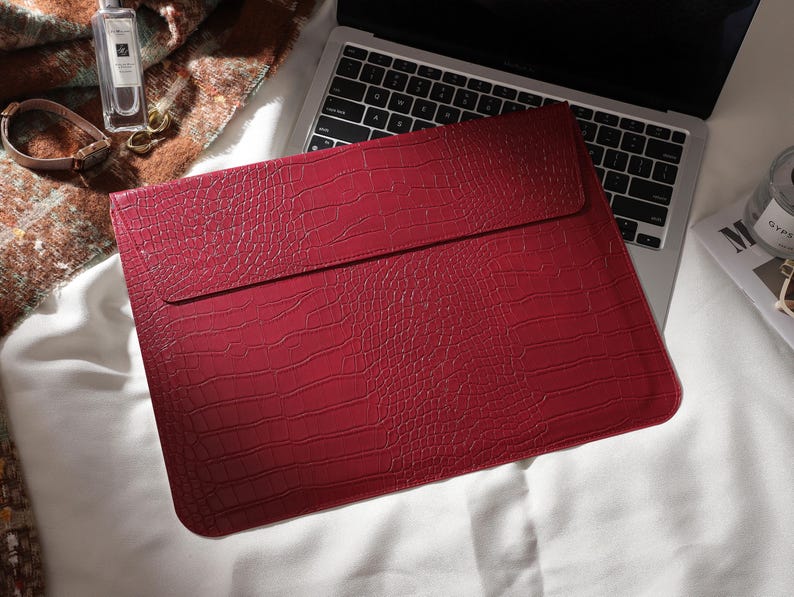 Puede incluir: Una funda roja para port&aacute;til con textura de piel de cocodrilo. La funda rectangular tiene un cierre de solapa. Est&aacute; sobre un teclado de port&aacute;til. El fondo incluye un frasco de perfume, un reloj y una manta.