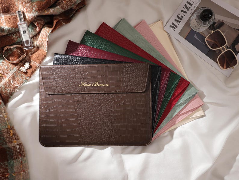 May include: A brown faux-leather laptop sleeve with a crocodile texture, featuring the name "Kaia Brown" in gold lettering. A fan of colorful laptop sleeves in various colors is displayed behind the brown sleeve. Accessories like a perfume bottle, sunglasses, and a magazine are also visible.