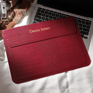 Puede incluir: Una funda roja para port&aacute;til con textura de piel de cocodrilo. El texto "Dania James" est&aacute; impreso en dorado. La funda est&aacute; sobre un teclado de port&aacute;til.
