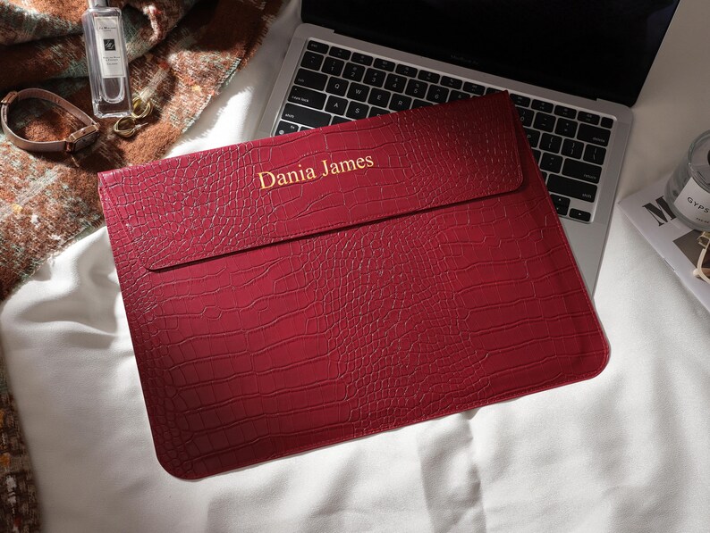Puede incluir: Una funda roja para port&aacute;til con textura de piel de cocodrilo. El texto "Dania James" est&aacute; impreso en dorado. La funda est&aacute; sobre un teclado de port&aacute;til.