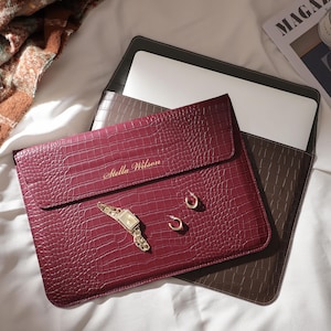 May include: A burgundy laptop sleeve with a crocodile texture, personalised with the name "Stella Wilson" in gold lettering. The sleeve is next to a brown laptop case, a gold watch, earrings, and a perfume bottle.