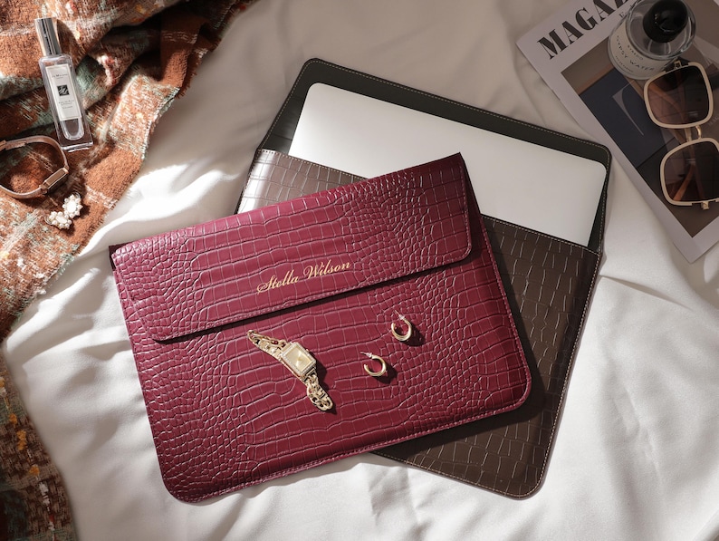 May include: A burgundy laptop sleeve with a crocodile texture, personalized with the name "Stella Wilson" in gold lettering. The sleeve is next to a brown laptop case, a gold watch, earrings, and a perfume bottle.