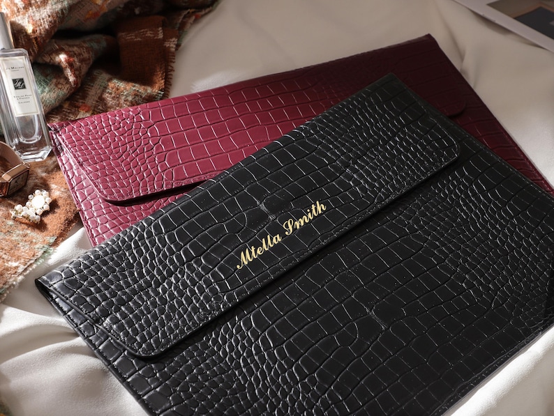 May include: A black faux-leather clutch with a crocodile texture, featuring the name "Miella Smith" in gold lettering. A burgundy clutch is visible behind it. The clutches are on a white surface.