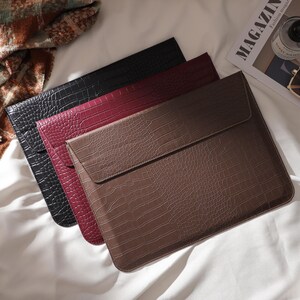 May include: Three faux-leather laptop sleeves in black, burgundy, and brown, arranged on a white surface. The sleeves have a crocodile skin texture and a flap closure. A magazine, sunglasses, and perfume bottle are also visible.