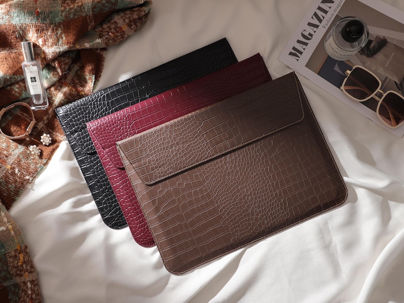 May include: Three faux-leather laptop sleeves in black, burgundy, and brown, arranged on a white surface. The sleeves have a crocodile skin texture and a flap closure. A magazine, sunglasses, and perfume bottle are also visible.