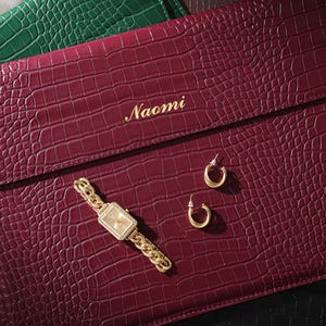 May include: A burgundy faux-leather laptop case with a crocodile texture, gold watch, and hoop earrings. The case is personalized with the name "Naomi" in gold lettering. A green case is visible in the background.