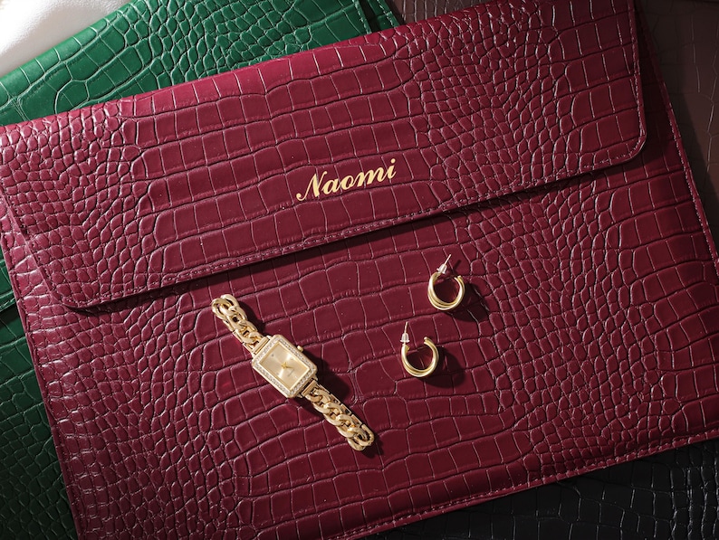 May include: A burgundy faux-leather laptop case with a crocodile texture, gold watch, and hoop earrings. The case is personalized with the name "Naomi" in gold lettering. A green case is visible in the background.