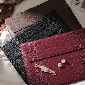 May include: An assortment of laptop sleeves in brown, black, and burgundy, featuring a crocodile skin texture. A gold wristwatch and pearl earrings are placed on the burgundy sleeve. The sleeves are arranged on a white surface.