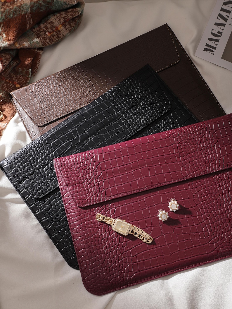 May include: An assortment of laptop sleeves in brown, black, and burgundy, featuring a crocodile skin texture. A gold wristwatch and pearl earrings are placed on the burgundy sleeve. The sleeves are arranged on a white surface.