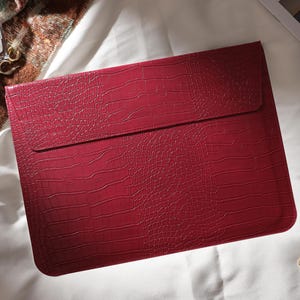 Puede incluir: Una funda para port&aacute;til burdeos con textura de piel de cocodrilo. La funda tiene un cierre de solapa y es rectangular. Tambi&eacute;n se ven un reloj de oro, pendientes y un frasco de perfume.