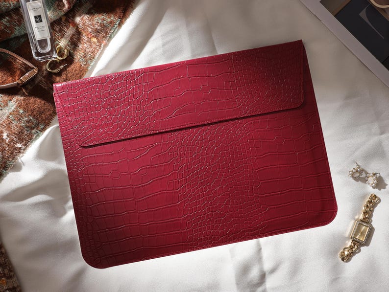 Puede incluir: Una funda para port&aacute;til burdeos con textura de piel de cocodrilo. La funda tiene un cierre de solapa y es rectangular. Tambi&eacute;n se ven un reloj de oro, pendientes y un frasco de perfume.