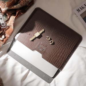 May include: A brown laptop sleeve with a crocodile skin texture, holding a silver laptop. A gold watch, earrings, and bracelet are on the sleeve. A perfume bottle, magazine, and sunglasses are nearby.