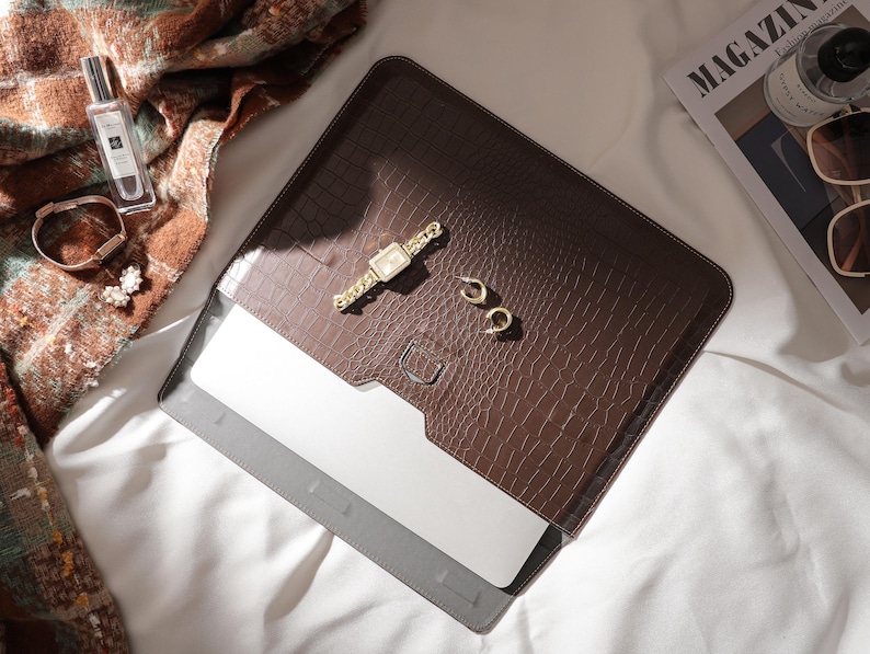 May include: A brown laptop sleeve with a crocodile skin texture, holding a silver laptop. A gold watch, earrings, and bracelet are on the sleeve. A perfume bottle, magazine, and sunglasses are nearby.