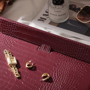 May include: A burgundy leather laptop case with a crocodile skin texture. A gold watch with a square face and a gold chain band, and two gold hoop earrings are on the case. A bottle of perfume, sunglasses, and a magazine are in the background.