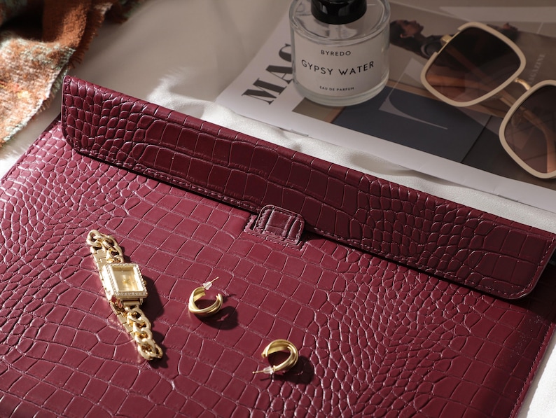 May include: A burgundy leather laptop case with a crocodile skin texture. A gold watch with a square face and a gold chain band, and two gold hoop earrings are on the case. A bottle of perfume, sunglasses, and a magazine are in the background.